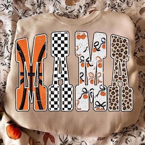 Puede incluir: Una sudadera beige con la palabra "MAMA" en letras grandes y audaces. La "M" es naranja con un dise&ntilde;o de baloncesto, la "A" es a cuadros blancos y negros, la segunda "M" es blanca con balones de baloncesto y cintas, y la &uacute;ltima "A" es estampado de leopardo.