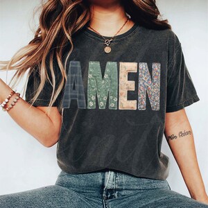 May include: Dark gray t-shirt with the word "AMEN" in patchwork letters. The letters feature plaid, floral, and patterned designs. The shirt is worn with blue jeans.
