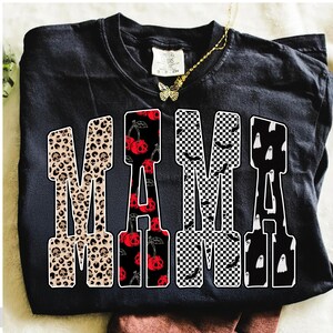 May include: Black t-shirt with the word "MAMA" in a bold, block letter design. The letters feature various patterns: leopard print with skulls, red cherries with skull faces, black and white checkered pattern with bats, and white ghosts.