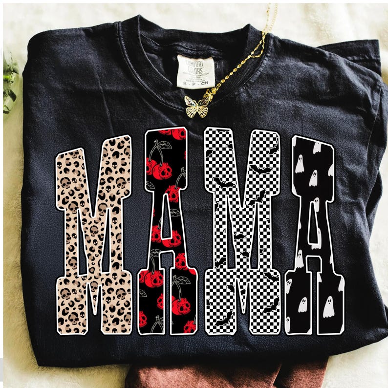 May include: Black t-shirt with the word "MAMA" in a bold, block letter design. The letters feature various patterns: leopard print with skulls, red cherries with skull faces, black and white checkered pattern with bats, and white ghosts.