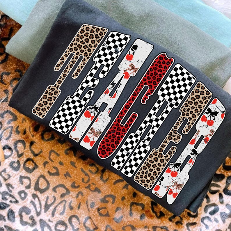 May include: Dark gray sweatshirt with the word "RADICAL" in a bold, stylized font. The letters feature a mix of patterns, including leopard print, checkered, and cherry designs. The sweatshirt is folded with other items in the background.