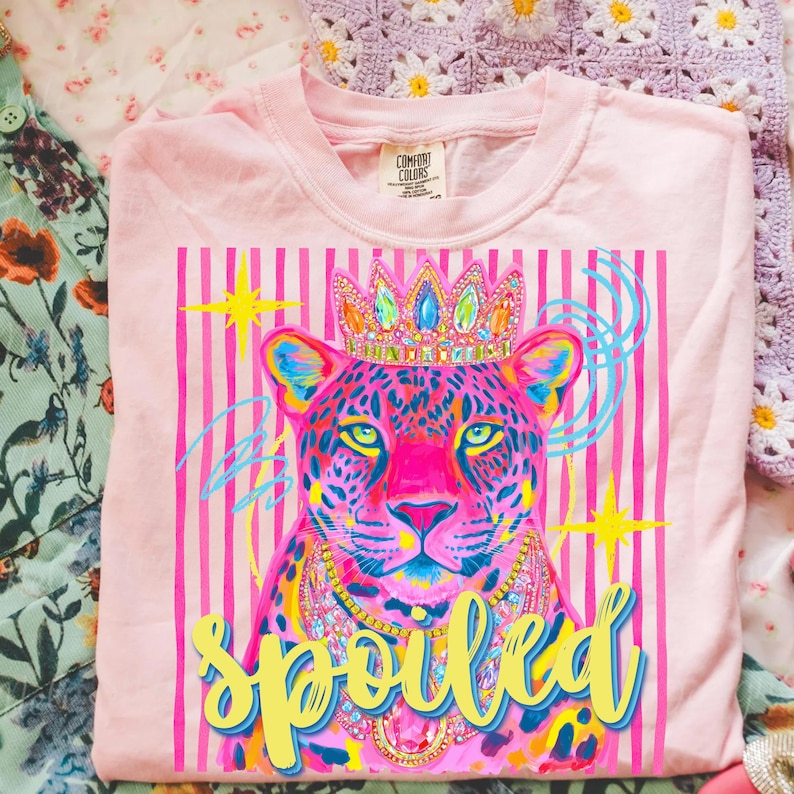 May include: A light pink t-shirt featuring a colorful illustration of a leopard wearing a crown and jewelry. The leopard is set against a pink striped background with the word "spoiled" in yellow script. The shirt has a Comfort Colors tag.
