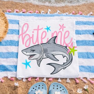 May include: White beach towel with a cartoon shark wearing a pearl necklace and the words "bite me" in pink script. The shark is surrounded by colorful starfish. The towel is on a sandy beach with blue and white striped fabric.