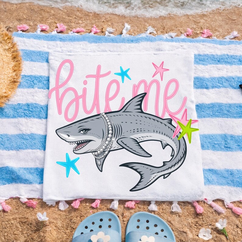 May include: White beach towel with a cartoon shark wearing a pearl necklace and the words "bite me" in pink script. The shark is surrounded by colorful starfish. The towel is on a sandy beach with blue and white striped fabric.