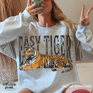 May include: A grey sweatshirt featuring the words "EASY TIGER" above a tiger illustration. The tiger is orange and black. The words "digital download" are at the bottom right.