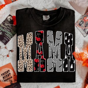 May include: Black t-shirt with the word "MAMA" in a bold, decorative font. The letters are filled with various Halloween-themed patterns, including leopard print skulls, cherries, checkered patterns with bats, and ghosts.