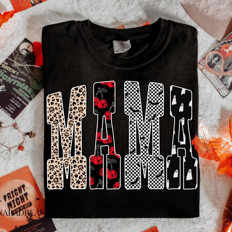 May include: Black t-shirt with the word "MAMA" in a bold, decorative font. The letters are filled with various Halloween-themed patterns, including leopard print skulls, cherries, checkered patterns with bats, and ghosts.