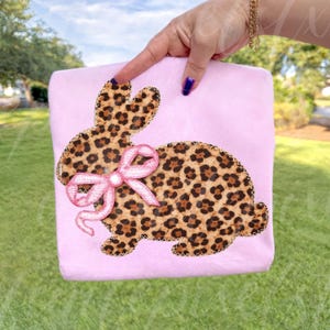 May include: A light pink, rectangular item featuring a leopard print bunny design with a pink bow. The bunny is outlined with a decorative stitch. The item is held by a hand with a gold bracelet and blue nail polish.