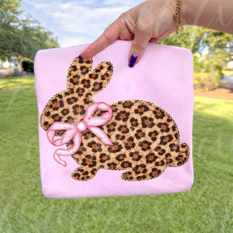 May include: A light pink, rectangular item featuring a leopard print bunny design with a pink bow. The bunny is outlined with a decorative stitch. The item is held by a hand with a gold bracelet and blue nail polish.