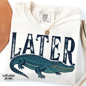 Pode incluir: Uma camiseta bege com um gr&aacute;fico desbotado de um jacar&eacute; e a palavra "LATER" em letras pretas.
