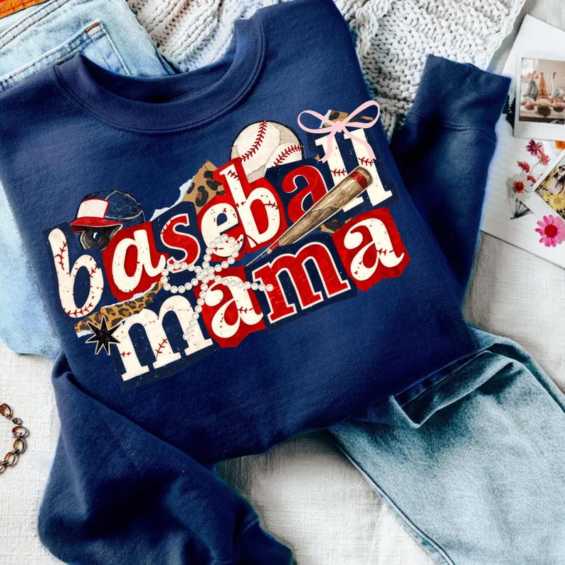 May include: Navy blue sweatshirt with the words "Baseball Mama" in a layered design. The design includes baseballs, a bat, a baseball cap, and a pearl necklace. The sweatshirt is surrounded by blue jeans and a light gray blanket.