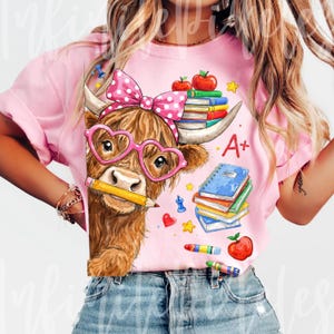 Pu&ograve; includere: T-shirt rosa con un cartone animato di una mucca delle Highlands con occhiali a forma di cuore e un fiocco a pois. La mucca tiene una matita, circondata da libri, mele e materiale scolastico. La maglietta ha la scritta "A+".