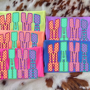 May include: A collection of six colorful t-shirts, each displaying the word "MAMA" in large, bold letters. The letters feature a checkered pattern in vibrant shades of pink, yellow, and blue. The shirts are in various colors, including yellow, orange, pink, purple, and blue.