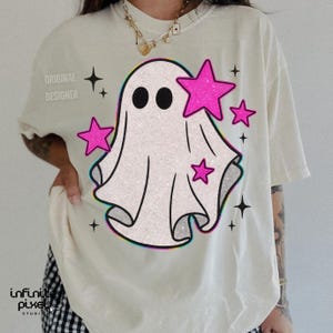 May include: Cream-colored t-shirt featuring a cartoon ghost with black eyes and pink stars. The ghost design has a glittery texture and a colorful outline. The words "ORIGINAL DESIGNER" are printed on the left shoulder.
