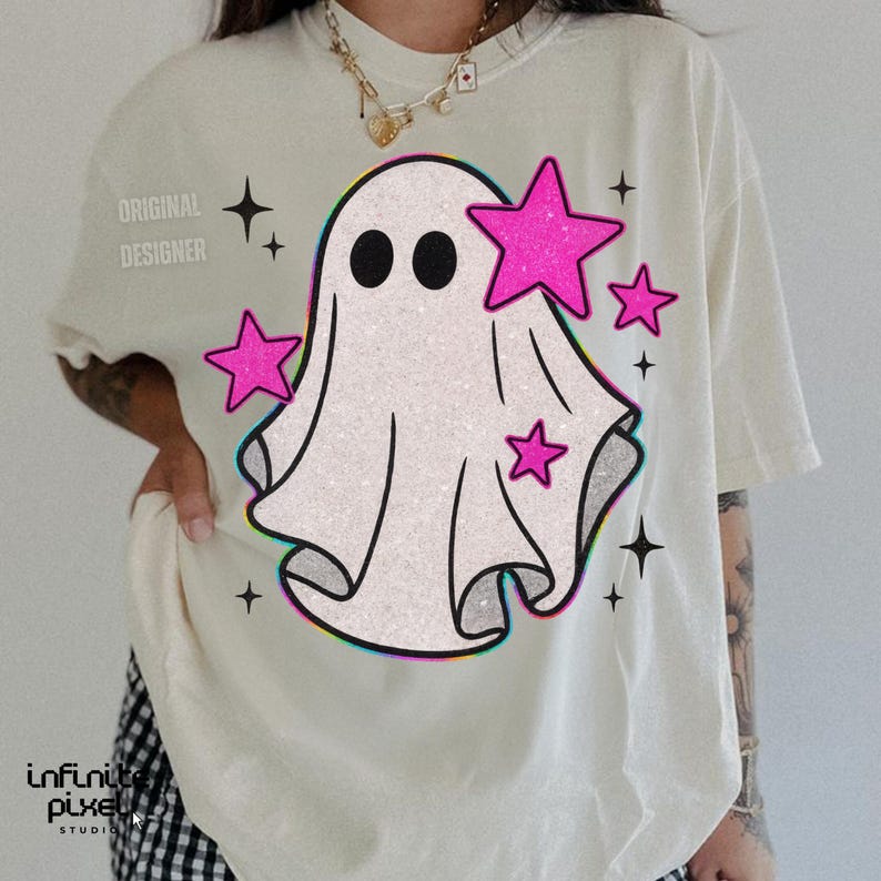 May include: Cream-colored t-shirt featuring a cartoon ghost with black eyes and pink stars. The ghost design has a glittery texture and a colorful outline. The words "ORIGINAL DESIGNER" are printed on the left shoulder.