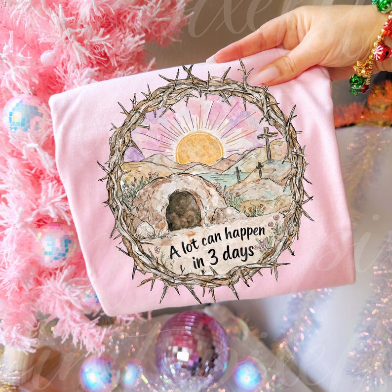 May include: A light pink t-shirt featuring a watercolor illustration of a sunrise over a landscape with three crosses and a tomb. The design is framed by a crown of thorns, with the text "A lot can happen in 3 days".