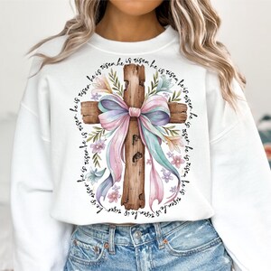 May include: White sweatshirt with a watercolor illustration of a wooden cross decorated with a pink and blue bow and floral accents. The text "He is risen" is repeated around the cross.