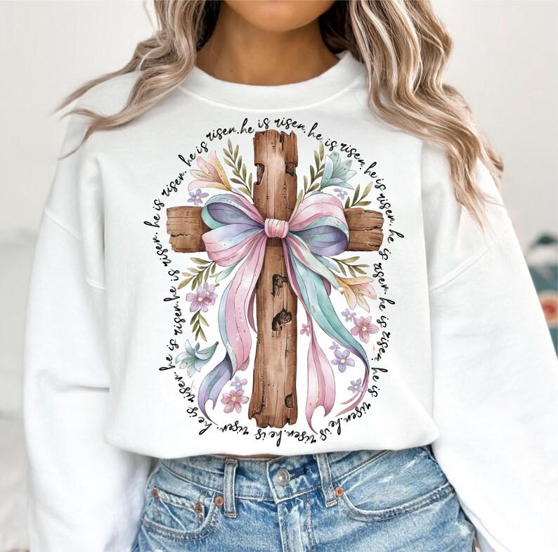 May include: White sweatshirt with a watercolor illustration of a wooden cross decorated with a pink and blue bow and floral accents. The text "He is risen" is repeated around the cross.