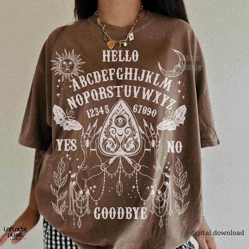 May include: Brown t-shirt featuring a white Ouija board design. The design includes the alphabet, numbers, sun, moon, butterflies, and the words "Hello", "Yes", "No", and "Goodbye". The shirt has short sleeves.