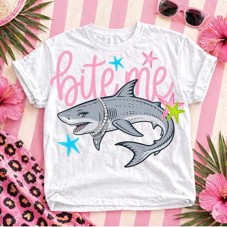 May include: White t-shirt with a graphic of a gray shark wearing a pearl necklace, the words "bite me" in pink script, and blue and green star accents. The shirt is displayed on a pink and white striped background.