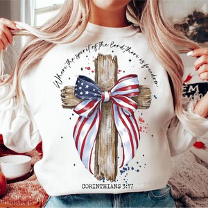 K&ouml;nnte beinhalten: Wei&szlig;es Sweatshirt mit einem Holzkreuz und einer rot-wei&szlig;-blauen Schleife. Das Kreuz ist mit Sternen und dem Text "Where the spirit of the Lord there is freedom" und "Corinthians 3:17" verziert.