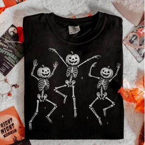 May include: Black t-shirt featuring a white graphic of three dancing skeletons with pumpkin heads. The skeletons are in a playful pose, arms raised, and legs bent. A Halloween-themed design.
