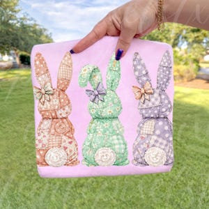 May include: A pink Easter-themed decorative pillow featuring three patchwork bunny rabbits. The bunnies are in shades of peach, green, and purple, each with a bow and a white, fluffy tail. The pillow is held by a hand.