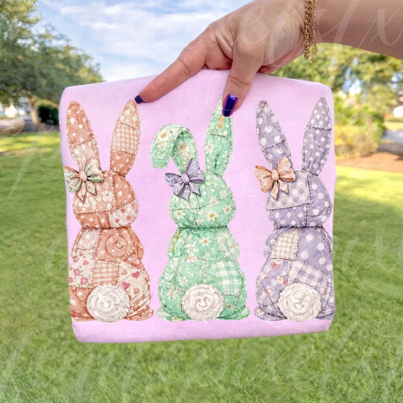May include: A pink Easter-themed decorative pillow featuring three patchwork bunny rabbits. The bunnies are in shades of peach, green, and purple, each with a bow and a white, fluffy tail. The pillow is held by a hand.