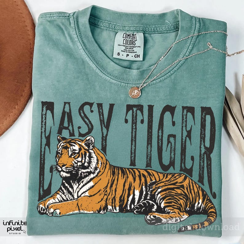 May include: A teal t-shirt with the text "EASY TIGER" in a distressed black font. The shirt features an illustration of a tiger in orange and black. A silver necklace with a pendant is draped on the shirt.