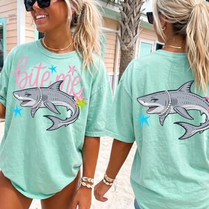 May include: Mint green t-shirt with a shark graphic wearing a pearl necklace. The front of the shirt has the text "Bite Me" in pink script. The shark is surrounded by stars in blue and yellow.