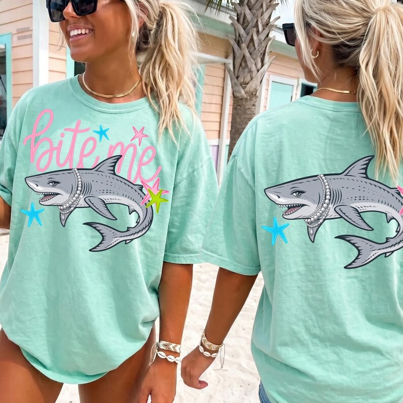 May include: Mint green t-shirt with a shark graphic wearing a pearl necklace. The front of the shirt has the text "Bite Me" in pink script. The shark is surrounded by stars in blue and yellow.