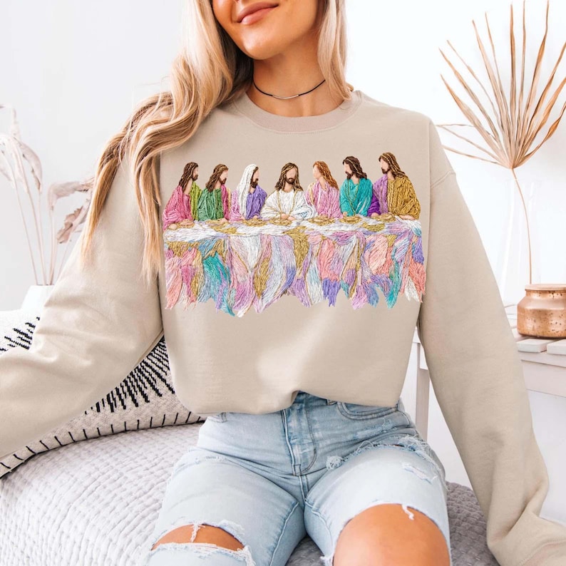 May include: Beige sweatshirt featuring an embroidered design of the Last Supper. The design includes figures in colorful robes seated at a table with a detailed, multi-colored tablecloth. The sweatshirt is a neutral color.