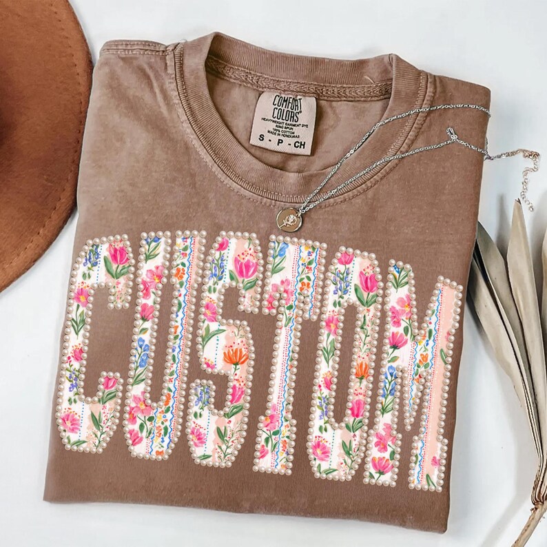 Puede incluir: Una camiseta marr&oacute;n Comfort Colors con la palabra "CUSTOM" en un dise&ntilde;o floral. Las letras est&aacute;n delineadas con perlas y rellenas de flores rosas, azules y naranjas. Un collar plateado con un colgante descansa sobre la camiseta.