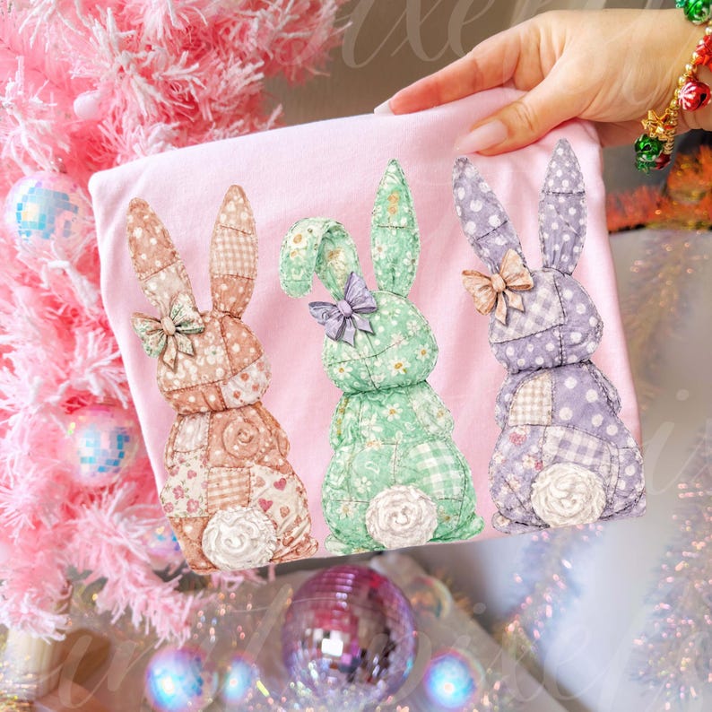 May include: A light pink t-shirt featuring three patchwork bunny rabbits in shades of peach, green, and purple. Each bunny has a bow and a fluffy white tail. The bunnies are set against a pink background.