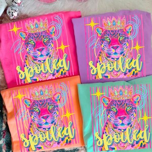 May include: Four colorful t-shirts with a leopard graphic wearing a crown and necklace. The word "spoiled" is written in yellow script below the leopard. The shirts are pink, purple, orange, and teal.