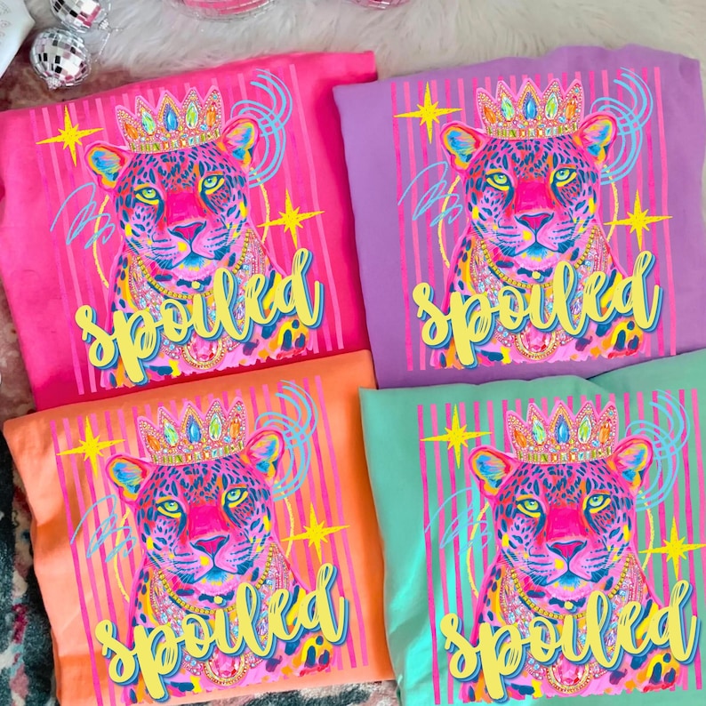 May include: Four colorful t-shirts with a leopard graphic wearing a crown and necklace. The word "spoiled" is written in yellow script below the leopard. The shirts are pink, purple, orange, and teal.