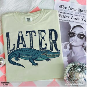 Pode incluir: Uma camiseta bege claro com um gr&aacute;fico de jacar&eacute; azul e a palavra "LATER" em uma fonte de estilo vintage.