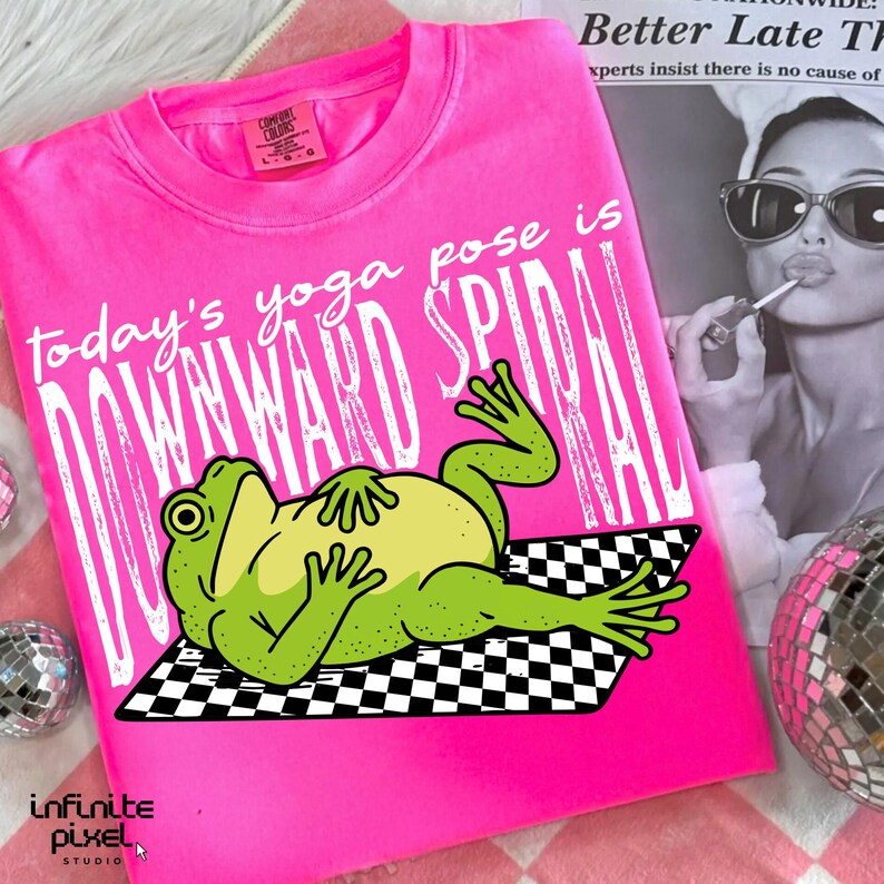以下が含まれることがあります： 鮮やかなピンク色のTシャツ。白文字で「today's yoga pose is DOWNWARD SPIRAL」と書かれています。ヨガのポーズをとるカエルのイラスト入り。Comfort Colorsブランド。