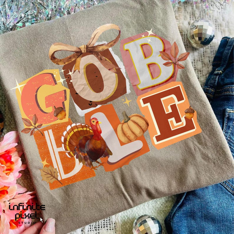 May include: A tan t-shirt featuring the word "GOBBLE" in colorful, block-letter style. The design includes a turkey, pumpkin, and fall leaves. A brown bow is at the top of the design. The shirt is folded on a surface with denim.