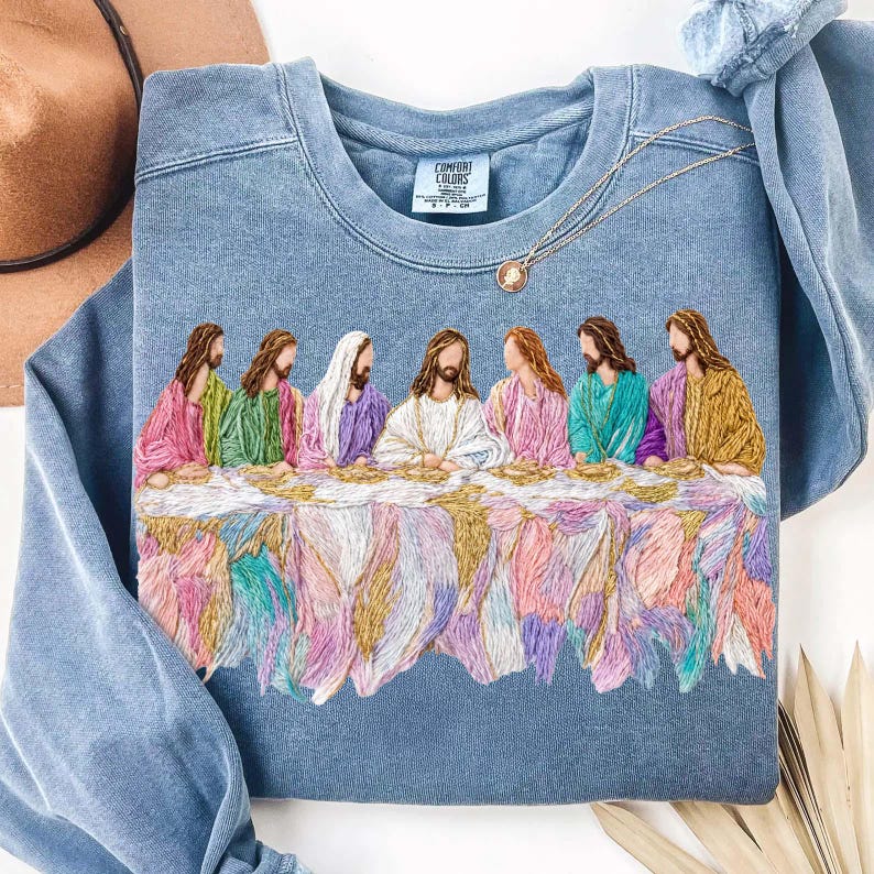 May include: A light blue crewneck sweatshirt featuring an embroidered depiction of the Last Supper. The embroidery showcases figures in various colors, including pink, green, and gold, seated around a table. A gold necklace with a pendant is draped on the sweatshirt.