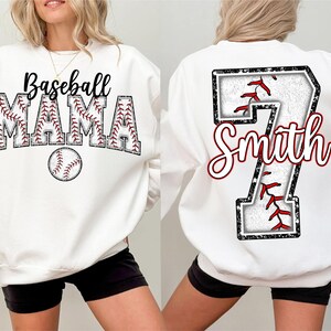 Pu&ograve; includere: Felpa bianca con un design a tema baseball. La parte anteriore presenta il testo "Baseball Mama" con una grafica di baseball. La parte posteriore presenta un grande numero "7" con il testo "Smith" e una grafica di baseball.