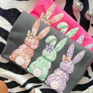 May include: Three decorative pillows with patchwork bunny designs in shades of pink, gray, and green. Each bunny has a bow and a floral or polka dot pattern. The pillows are on a zebra-striped surface.