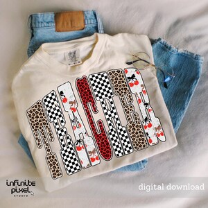 May include: Cream-colored t-shirt with the word "TEACHER" in a unique design. The letters feature leopard print, checkered patterns, and cherry illustrations. The shirt is laid on top of blue jeans.