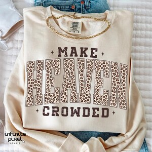 May include: A cream-colored sweatshirt with the text "MAKE HEAVEN CROWDED" in brown. The word "HEAVEN" is filled with a leopard print pattern. A gold chain necklace is draped across the top of the sweatshirt.