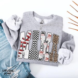 May include: A grey sweatshirt with the word "MAMA" in a decorative font. The letters feature baseballs, a black and white chequered pattern, baseballs with bows, and a leopard print design. A silver necklace is visible.