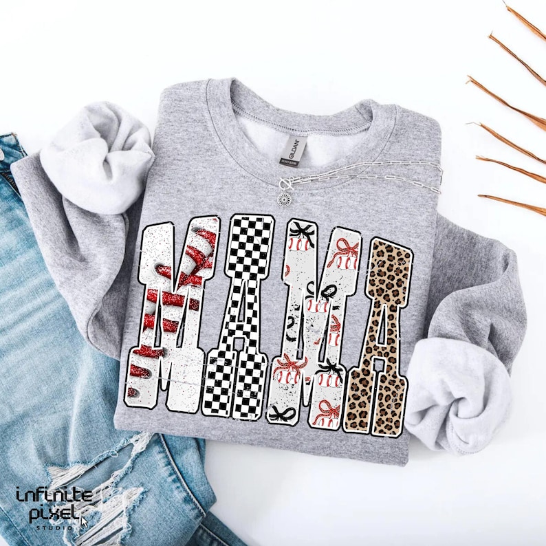 May include: A grey sweatshirt with the word "MAMA" in a decorative font. The letters feature baseballs, a black and white chequered pattern, baseballs with bows, and a leopard print design. A silver necklace is visible.