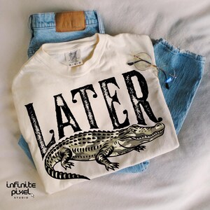 Peut inclure: T-shirt cr&egrave;me avec le mot "LATER" en grosses lettres noires et une illustration d'alligator. Le t-shirt est pos&eacute; sur un jean bleu clair, avec des lunettes &agrave; monture ronde.