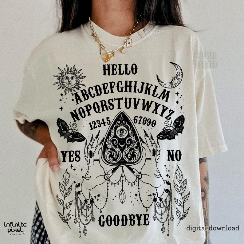 May include: Cream t-shirt featuring a black Ouija board design. The graphic includes the alphabet, numbers, and the words "Hello," "Yes," "No," and "Goodbye." Decorative elements include a sun, moon, butterflies, and hands.
