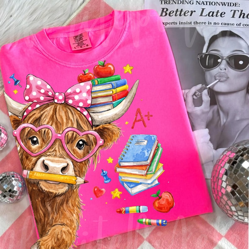 Pu&ograve; includere: Una maglietta rosa acceso con una mucca delle Highlands a cartoni animati che indossa occhiali a forma di cuore e un fiocco a pois. La mucca tiene una matita in bocca ed &egrave; circondata da elementi grafici a tema scolastico, tra cui libri, mele e la scritta "A+".
