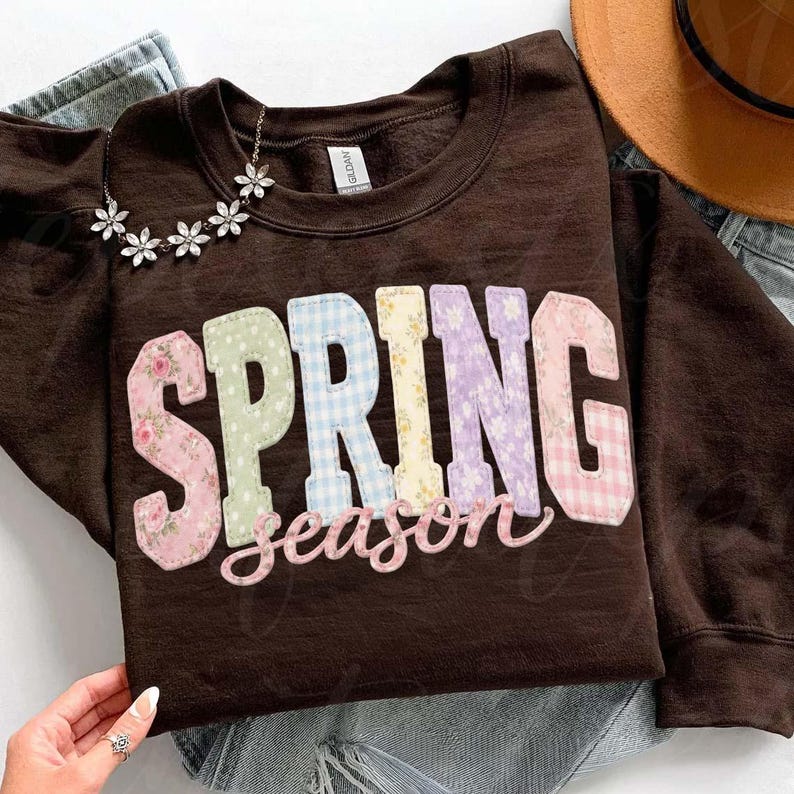 May include: A brown sweatshirt with the words "SPRING season" in colorful, patchwork-style letters. The letters are in various pastel patterns, including floral and gingham. A silver flower necklace is draped on the sweatshirt.